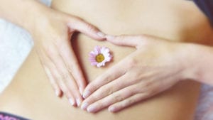 A woman makes a heart shape over her belly, where a single flower rests. The Microbiome Labs MegaSporeBiotic and other products may help you love your microbiome again!